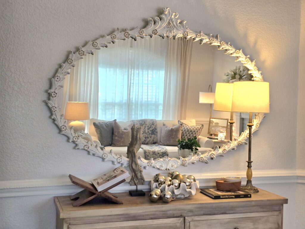 Carved white mirror above a wood console table styled with lamps, books, and decorative accents in warm lighting.