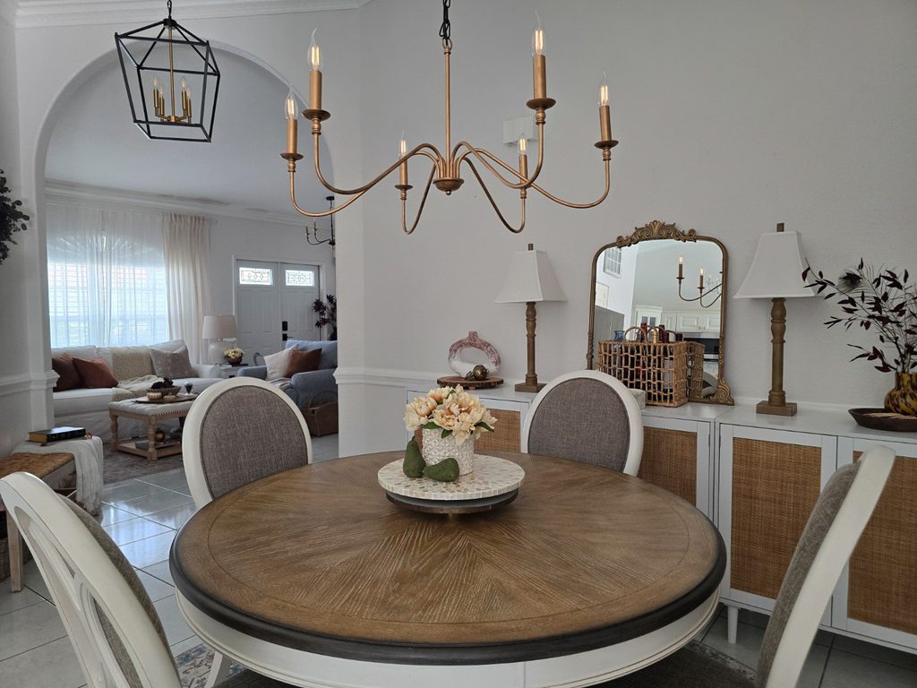 Round wood dining table beneath a brass chandelier with an arched opening into the living room.