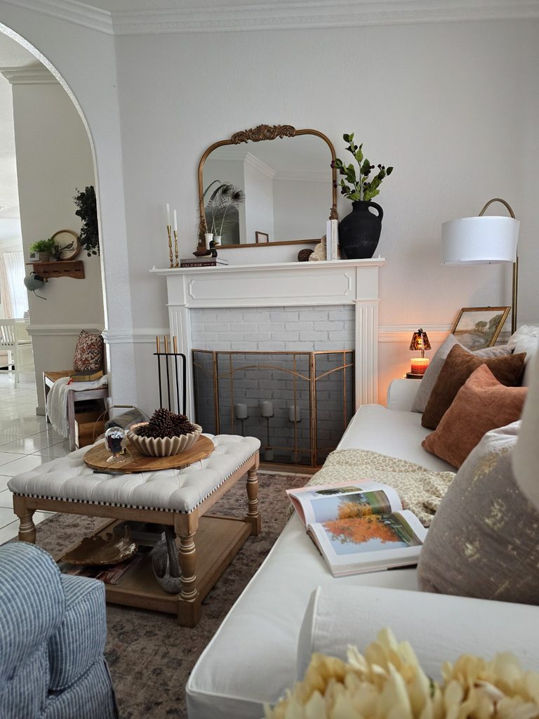 White sofa with open book, warm throw pillows, fireplace, coffee table and gold mirror.
