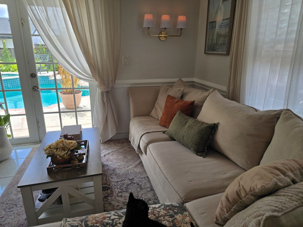 Family room sofa with green and caramel pillows, French doors, brass sconces and bird wall decor.