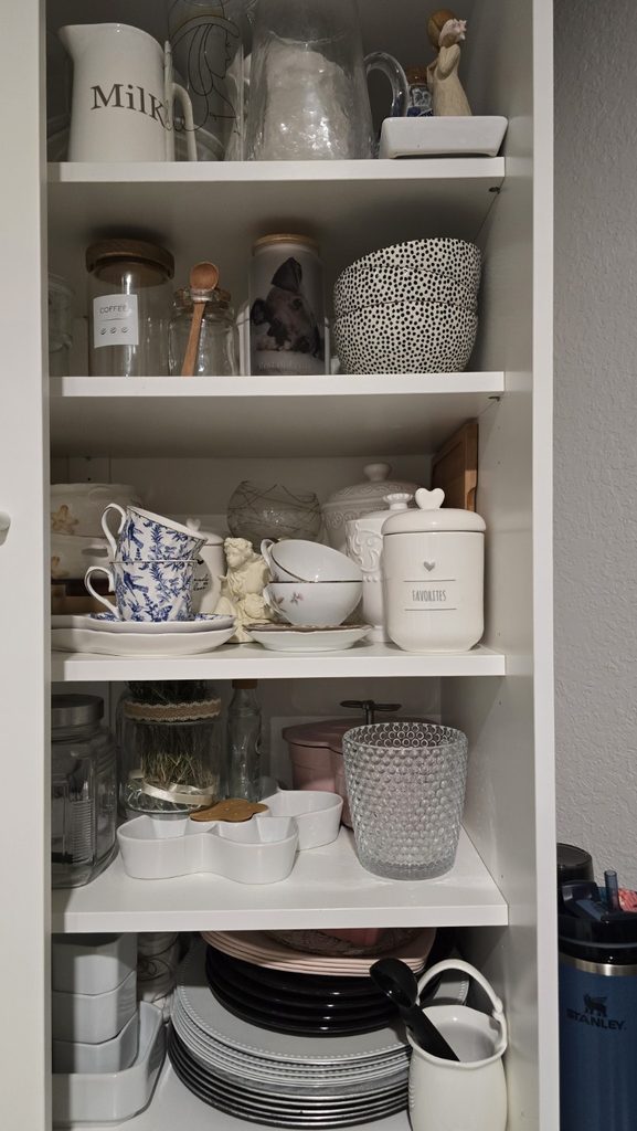 Open pantry shelves with pitchers, bowls, blue-and-white cups, ceramics, and stacked dishes.