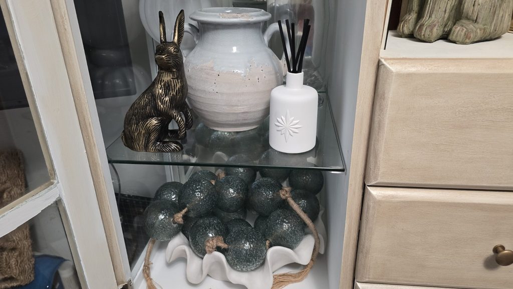 Cabinet shelves with a brass rabbit, ceramic vessel, diffuser, and dark green glass orbs.