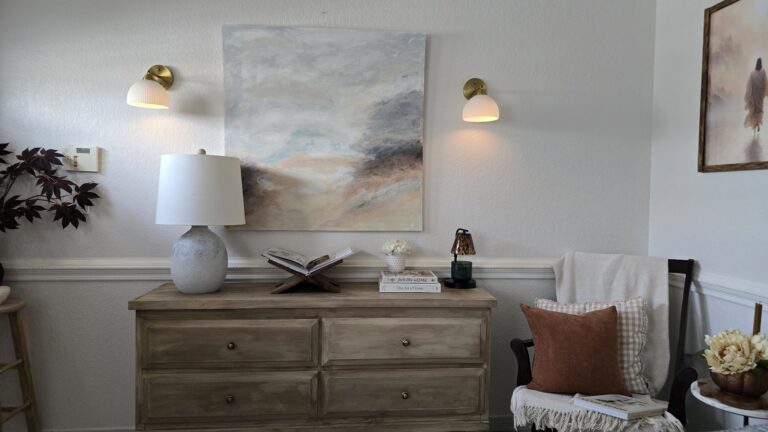 A wooden dresser styled with a large table lamp, open book, small lamp, and stacked books, beneath abstract art flanked by two brass sconces, with an armchair beside it.