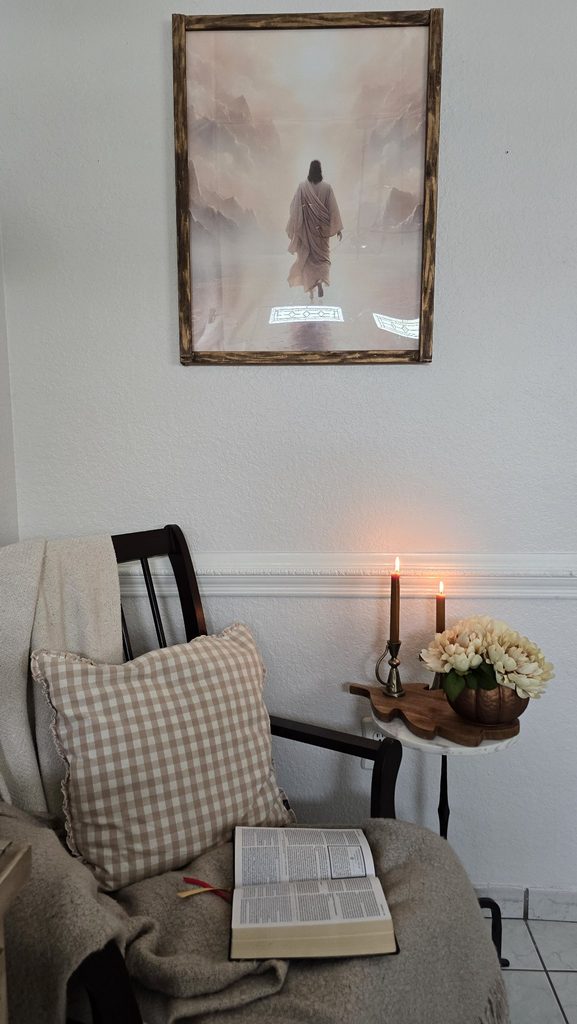 Reading corner: open Bible on a chair with gingham pillow and throw, lit candles, white flowers, framed painting of Jesus walking on water above.