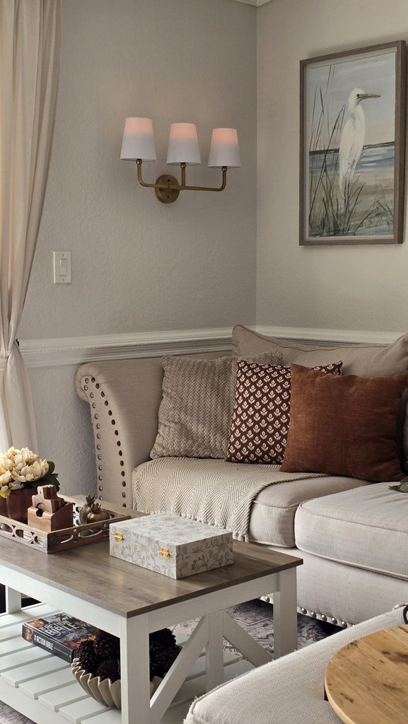 A sofa corner with a brass wall sconce, framed egret artwork, patterned and solid throw pillows, and a coffee table with a tray and books.