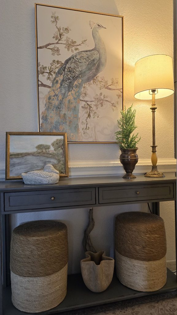 A console table styled with large peacock artwork, a small gilt-framed landscape, a white duck figurine, a brass urn with greenery, and a tall lamp, with woven stools below.