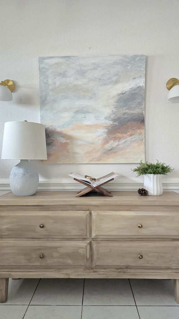 Styled dresser: lit lamp, open book on a wooden stand, white vase with greenery, a pinecone, landscape painting above.
