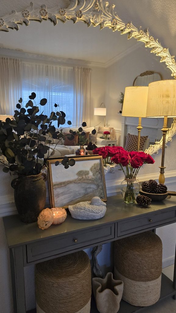 Console at dusk: lit table lamp, red roses in a glass vase, bowl of pinecones, mirror reflecting the lamplit living room.