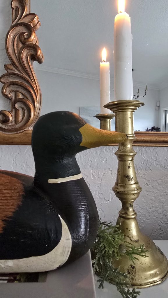 Aged brass candlestick with patina, lit candle reflected in a mirror, beside a wooden duck decoy.